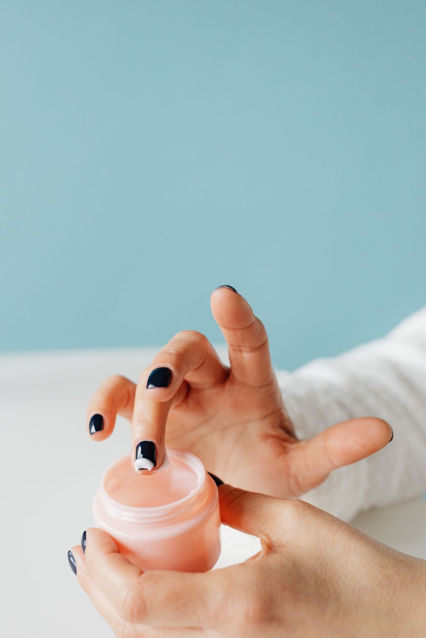 A close-up of hands applying moisturizing skincare cream from a pink jar against a light blue background.