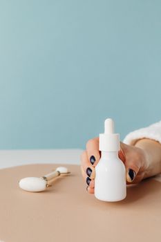 A close-up of a hand holding a skincare dropper bottle with a jade roller on a pastel backdrop.