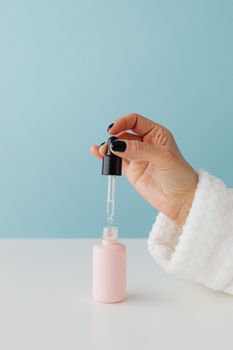 Close-up of a hand with black nails using a glass dropper in a pink bottle against a light blue backdrop.