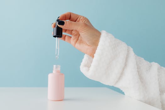 Close-up of a woman's hand in a bathrobe using a serum dropper over a pink glass bottle against a blue background.