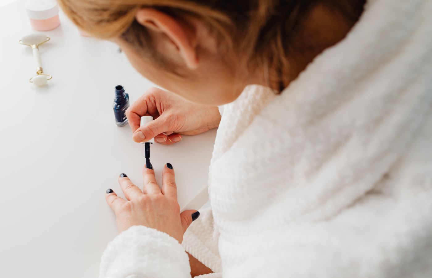 A woman in a bathrobe paints her nails indoors as part of a self-care routine.