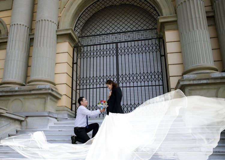 Man Proposing On Stairs With Flowers To Woman In White Dress Flowing In Wind