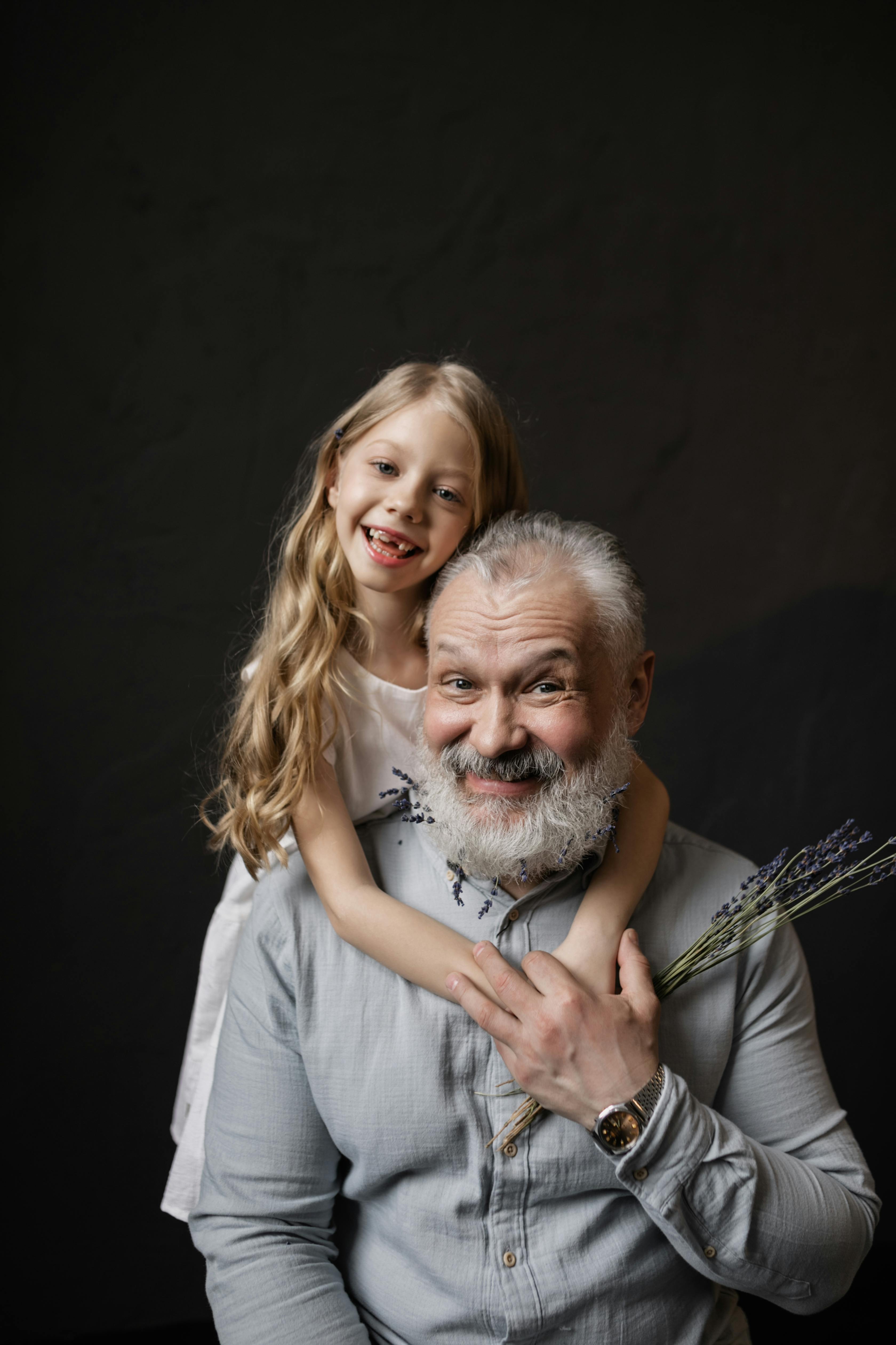 Blonde girl lovingly embracing her smiling bearded grandfather in a studio setting, capturing a heartwarming bond.
