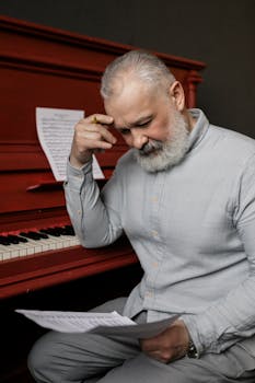 A mature composer thoughtfully examines sheet music next to a red piano, indicating deep focus and creativity.