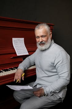 Elderly man with a beard composing music at a red piano, focused on his work.