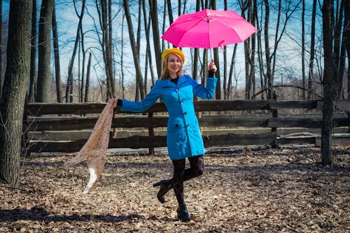 Full length of cheerful young female with umbrella and kerchief standing in autumn park while looking at camera