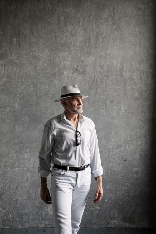 Free Stylish elderly man posing in a studio with a fedora hat, wearing white shirt and pants, looking away. Stock Photo