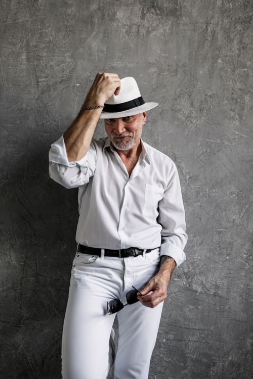 Free A confident senior man in white attire and fedora posing in a studio setting. Stock Photo