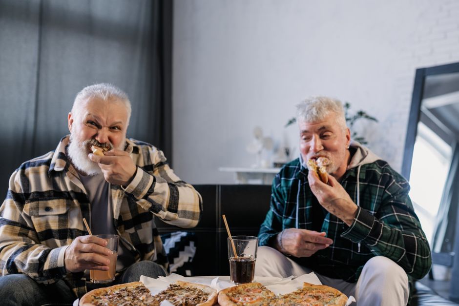 Two senior men having fun, eating pizza, and drinking cola indoors.