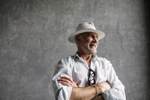 Free Elderly man smiling with folded arms, wearing a white fedora in a studio setting. Stock Photo