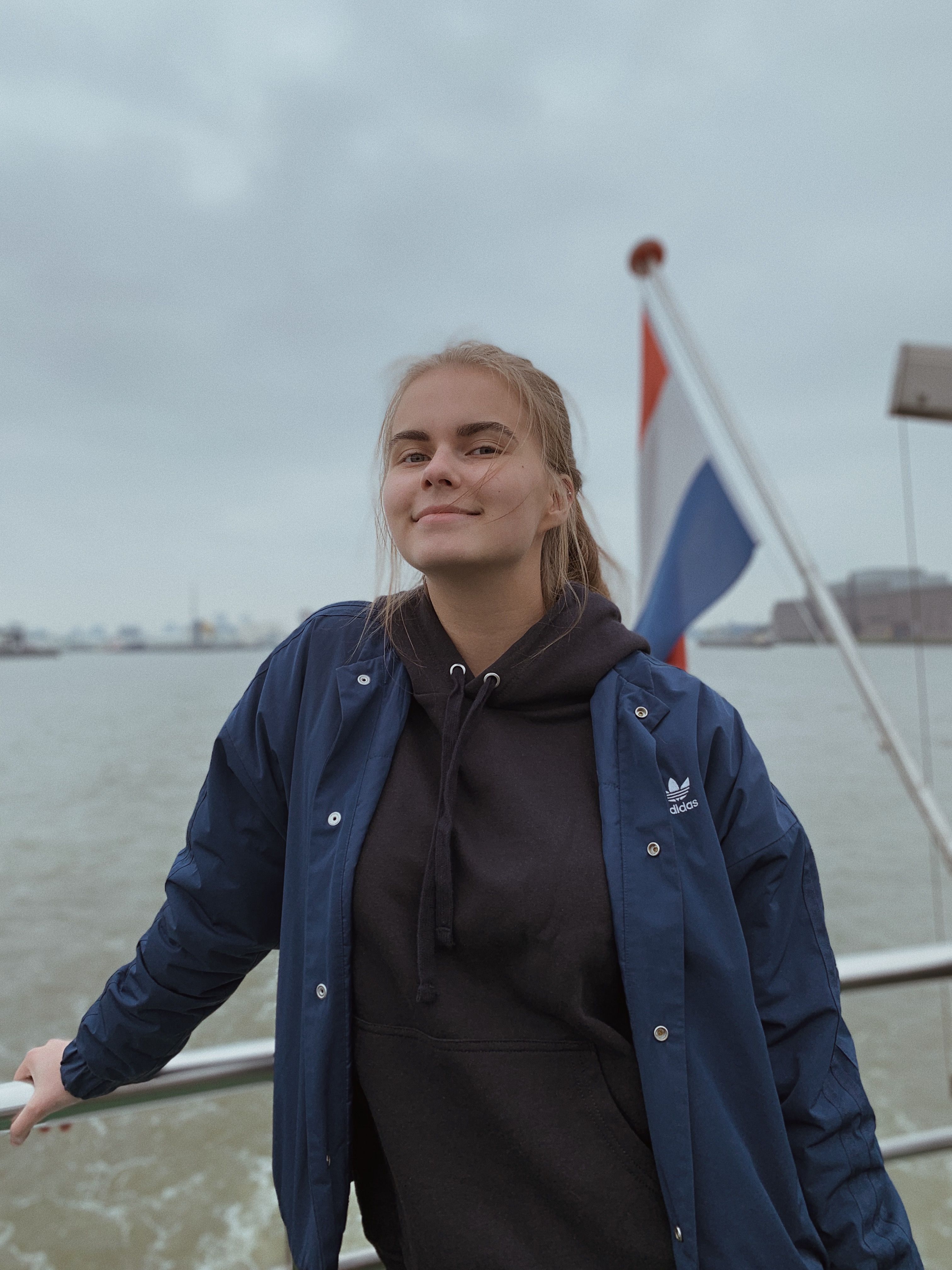 Smiling woman in a blue jacket poses on a boat in Rotterdam harbor with a Dutch flag in the background.