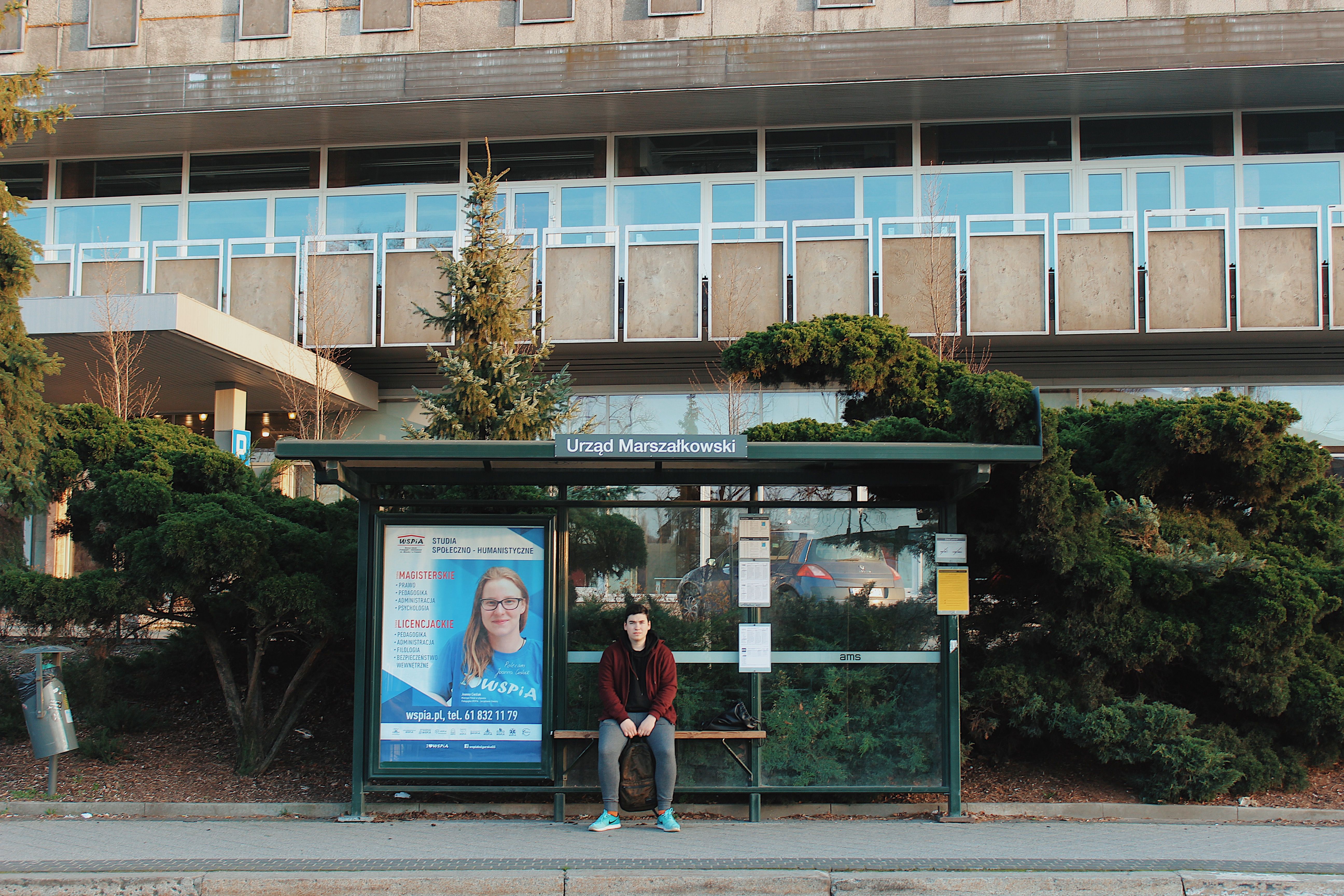 A man sitting at a bus stop in front of a modern building with greenery.