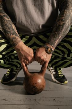 Close-up of a tattooed man in workout gear lifting a kettlebell indoors, focusing on strength and fitness.