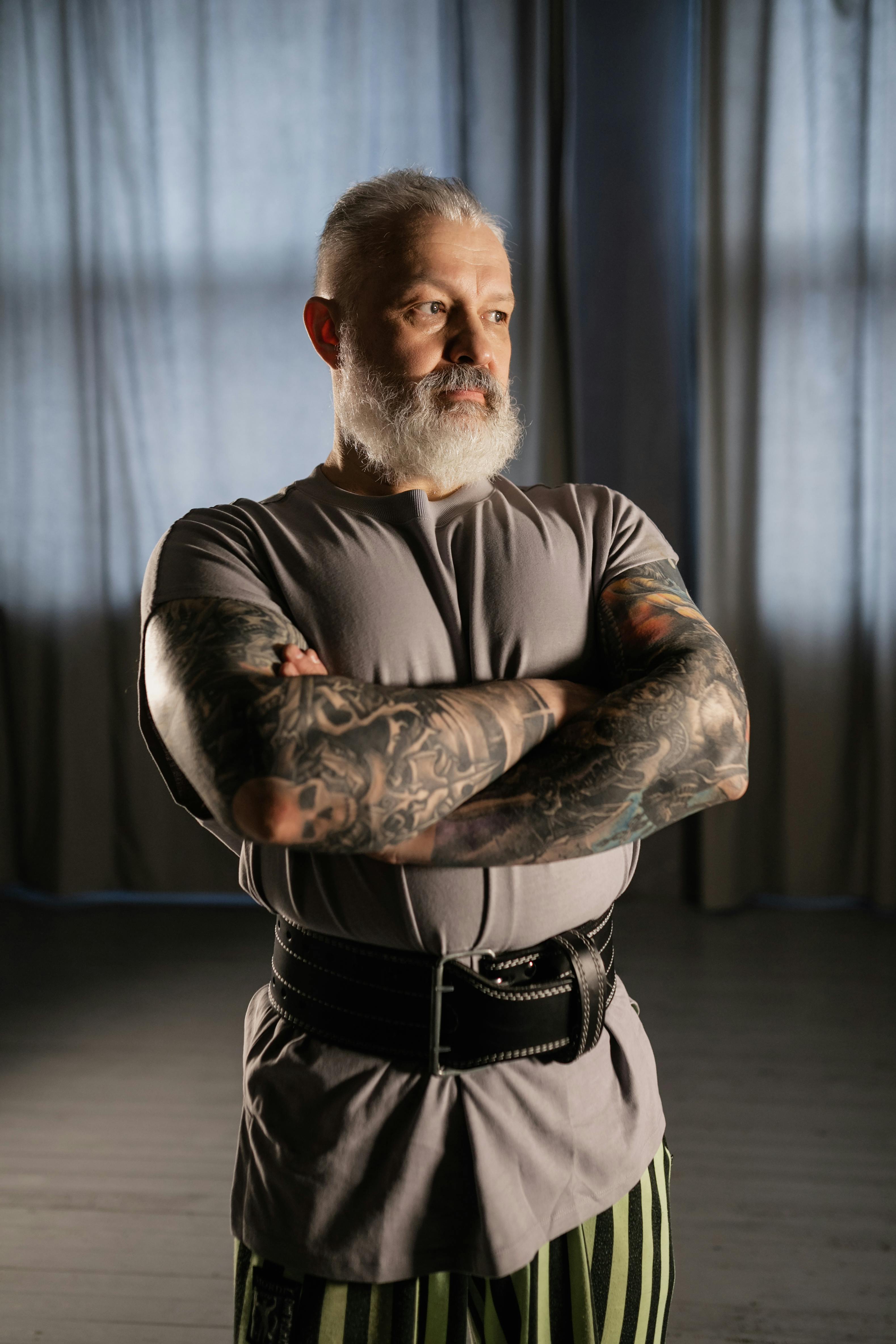 Elderly man with tattoos and a beard standing confidently indoors.