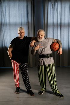 Two senior men in patterned pants pose confidently with a basketball indoors, showcasing positive aging.