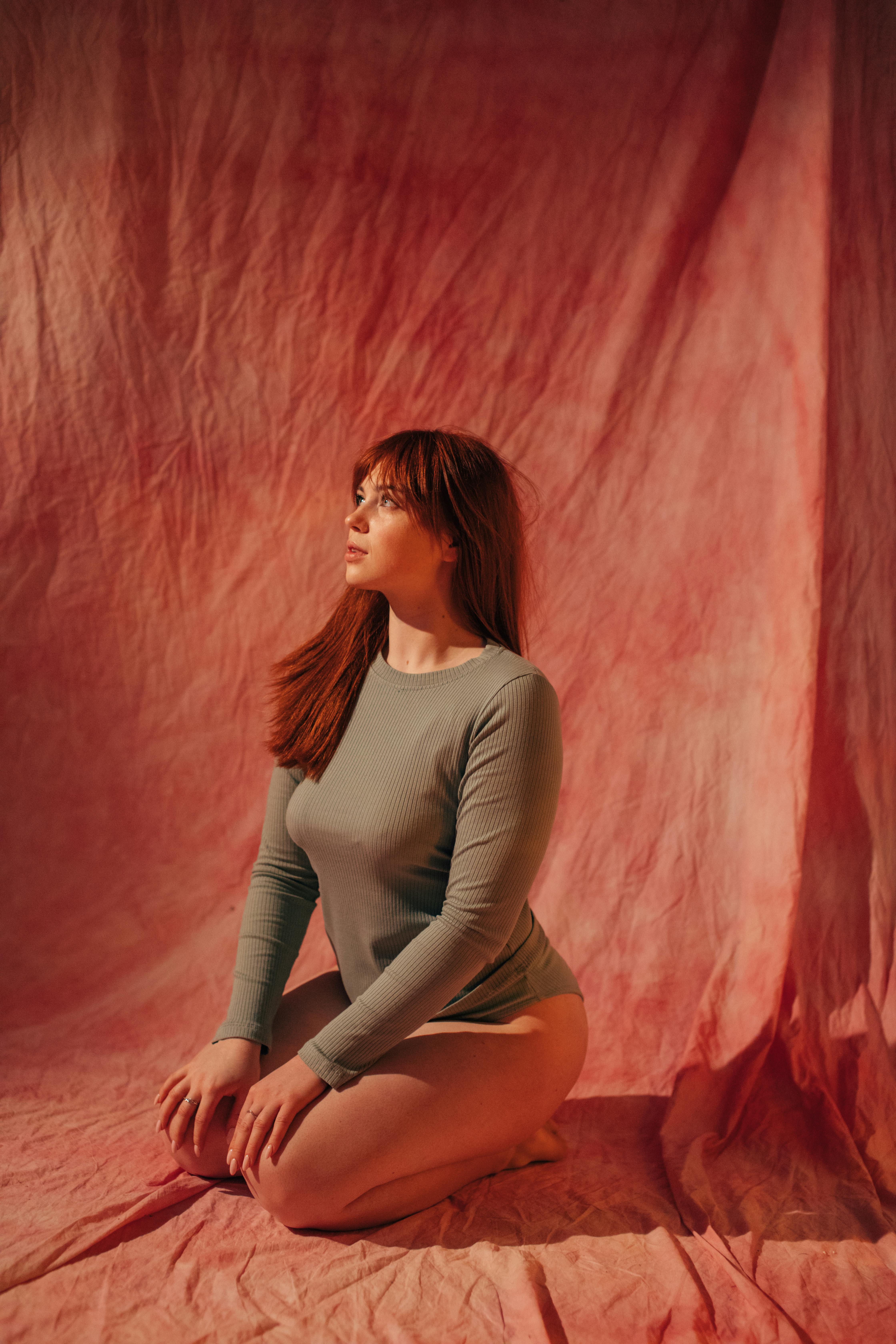 红头发的女人坐在粉红色的布料上，穿着绿色的长袖，若有所思地摆姿势