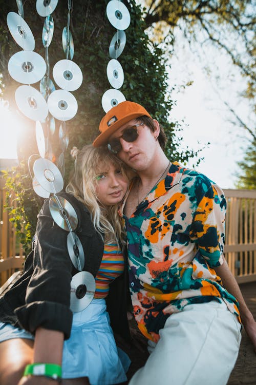 Free Young trendy male and female best friends in stylish outfits sitting on bench and cuddling near vintage music discs hanging from tree Stock Photo