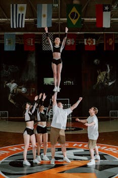 Cheerleaders performing an impressive stunt on an indoor court with flags overhead.