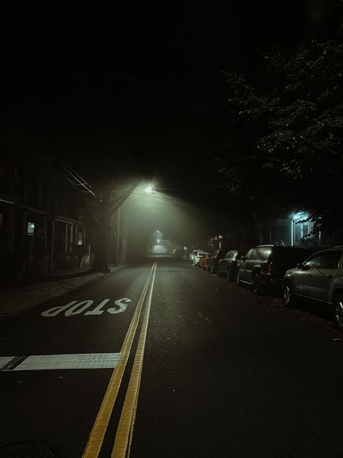 Free A misty urban street at night with parked cars and streetlights casting a moody atmosphere. Stock Photo
