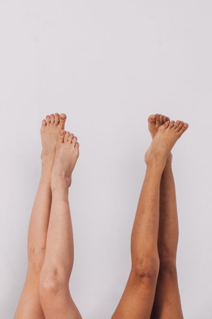 Two pairs of diverse legs raised barefoot against a white wall in a minimalist setting.
