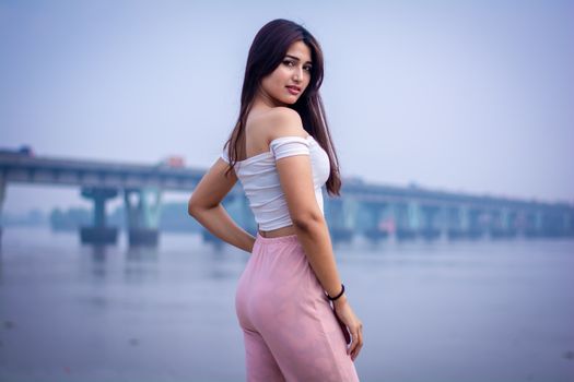 Confident young woman posing by a riverside with a bridge in the background.