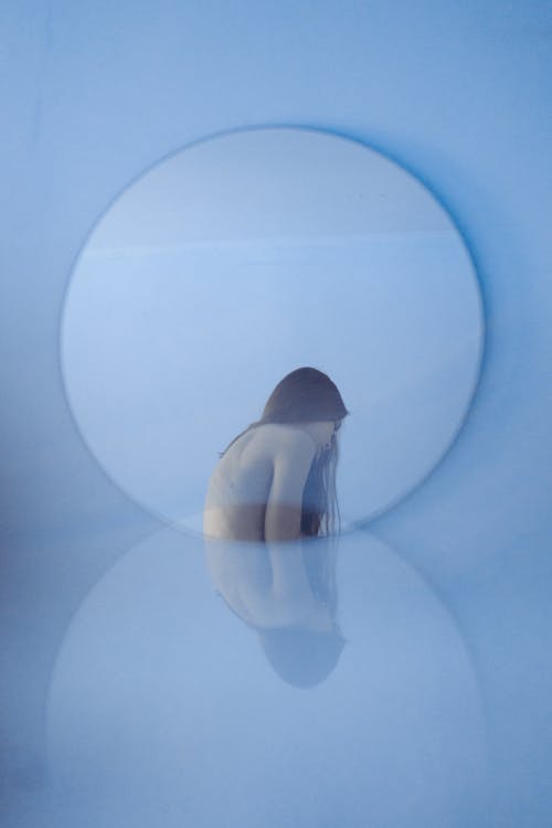 Free Side view of lonely woman with brown hair and tattoo on back against round shaped mirror Stock Photo