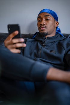 Young man in blue durag using smartphone while sitting on a sofa indoors.