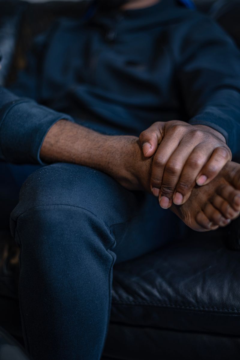 A Black man seated on a couch holds his bare foot with both hands, the posture people take when heel and arch pain flares.