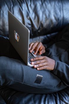 Hands of a person typing on a laptop while relaxed on a black leather sofa.