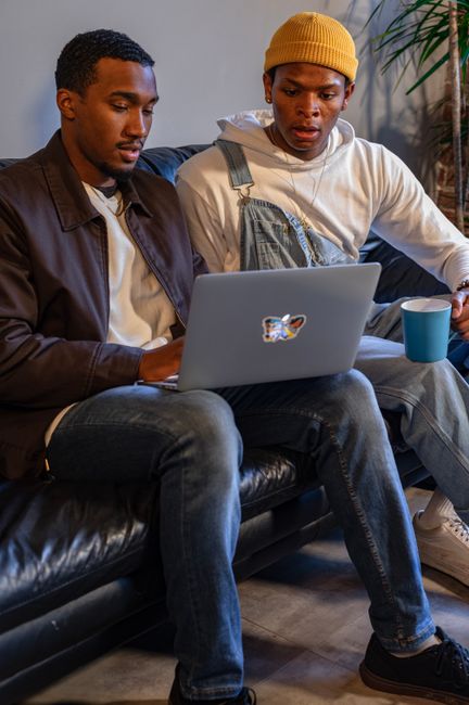 Two friends sitting on a sofa, working together on a laptop in a cozy indoor setting. Two friends sitting on a sofa, working together on a laptop in a cozy indoor setting.