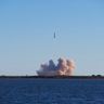 A rocket launches into a bright sky, leaving a thick trail of white smoke over the ocean.