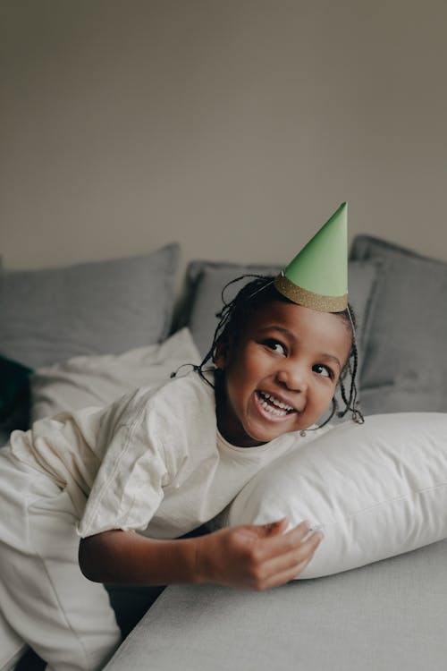 Little Girl in a Birthday Hat Standing next to a Sofa and Smiling 