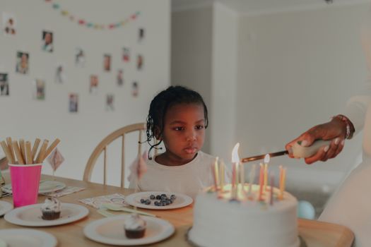 Young girl anticipating her birthday cake as an adult lights candles at indoor celebration.