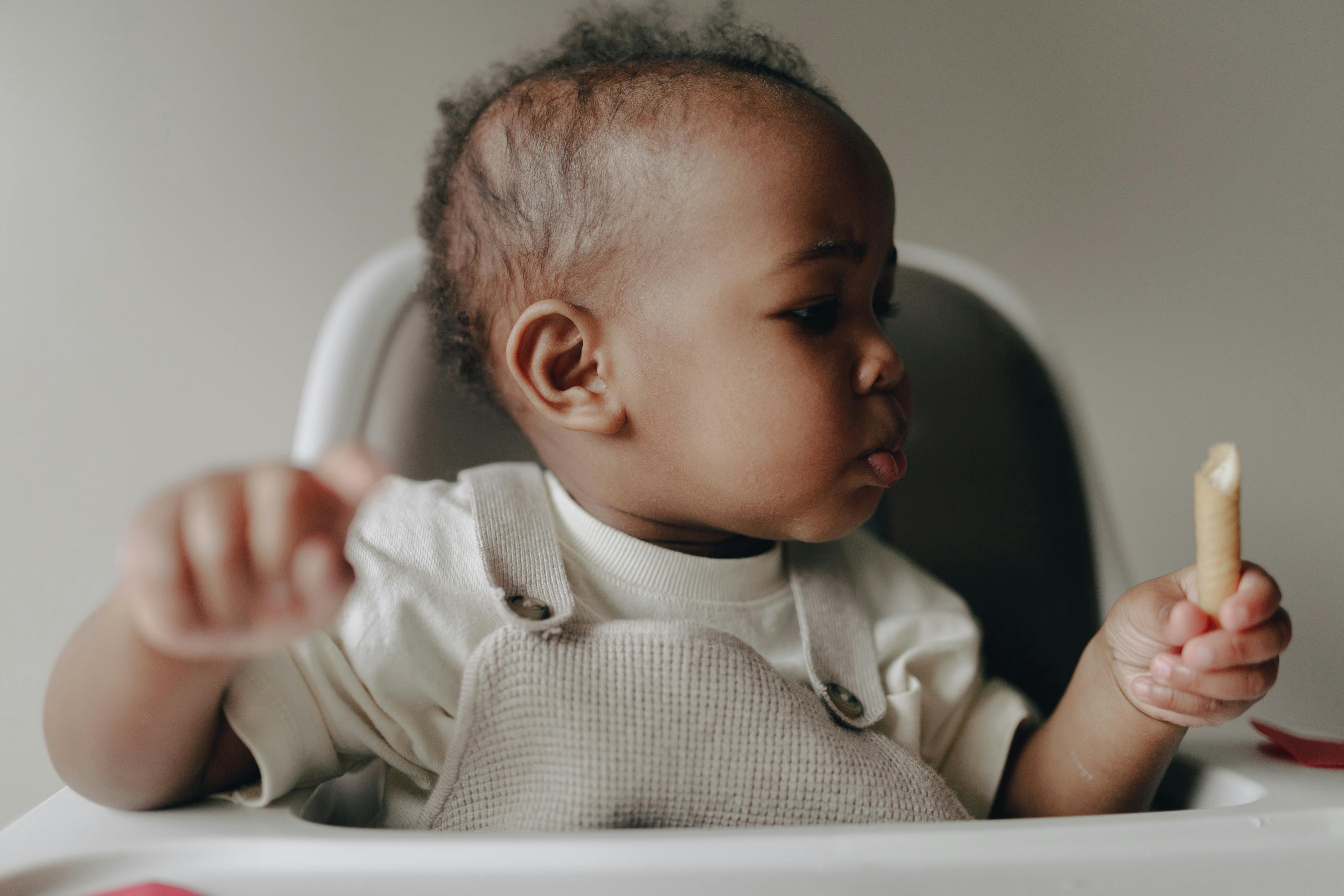 Cute baby sitting in high chair, enjoying a biscuit, with a curious facial expression.