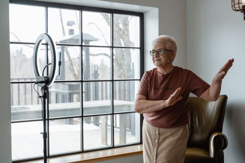 Free Elderly woman joyfully dancing indoors near a large window with ring light. Stock Photo
