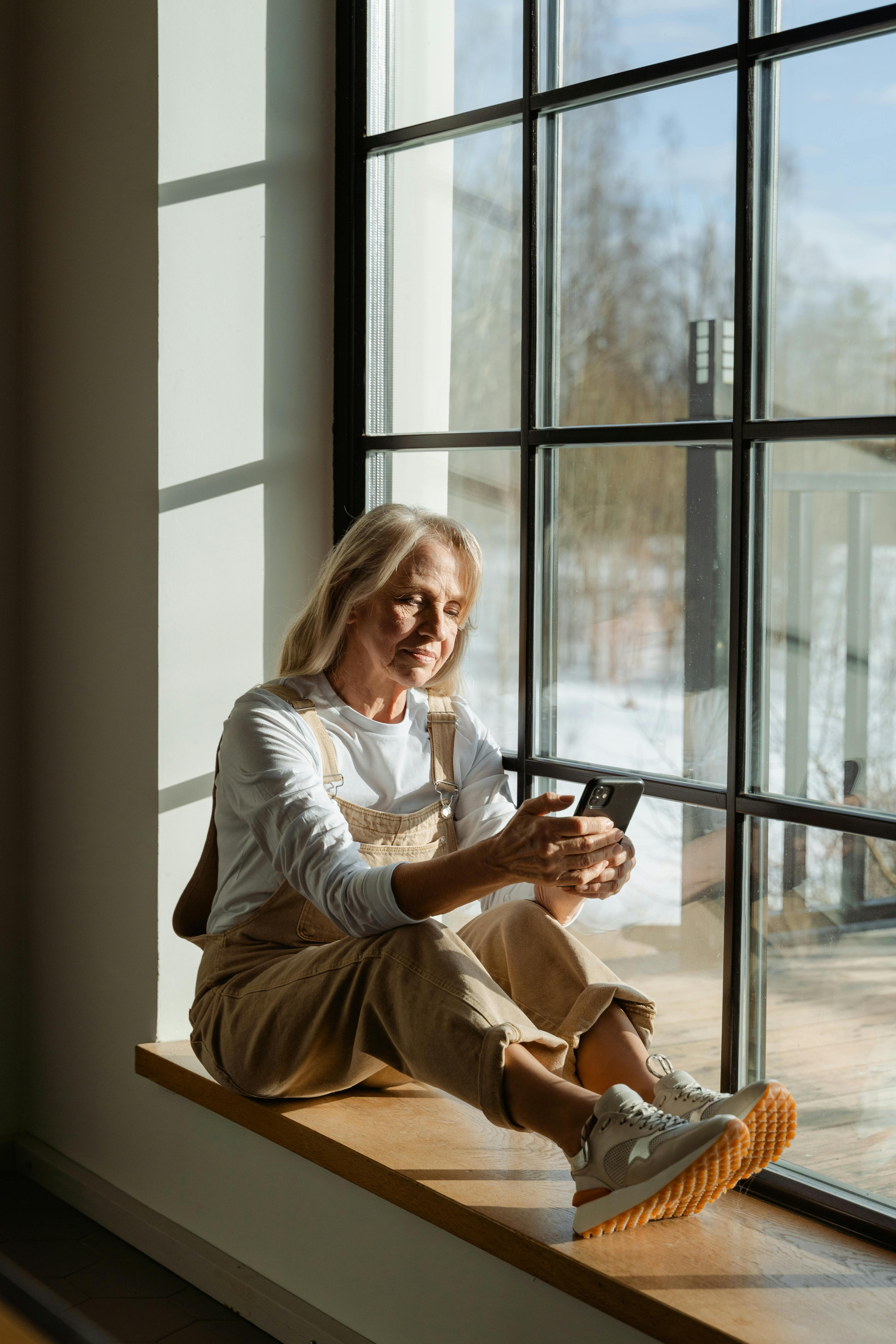 A senior woman using a smartphone while sitting by a window, enjoying the sunlight.