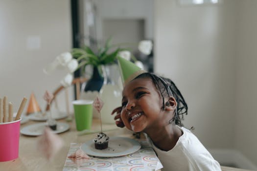A smiling child at a birthday party indoors, wearing a green party hat and enjoying the celebration.