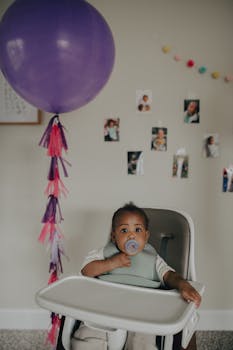 Cute toddler sitting in a high chair with purple balloon for a home celebration.