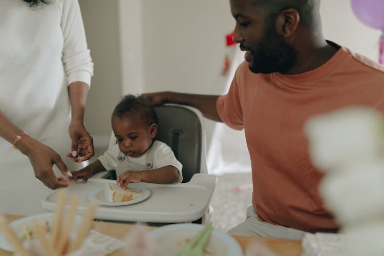 Mother And Father Sitting Next To A Baby Eating A Slice Of Birthday Cake