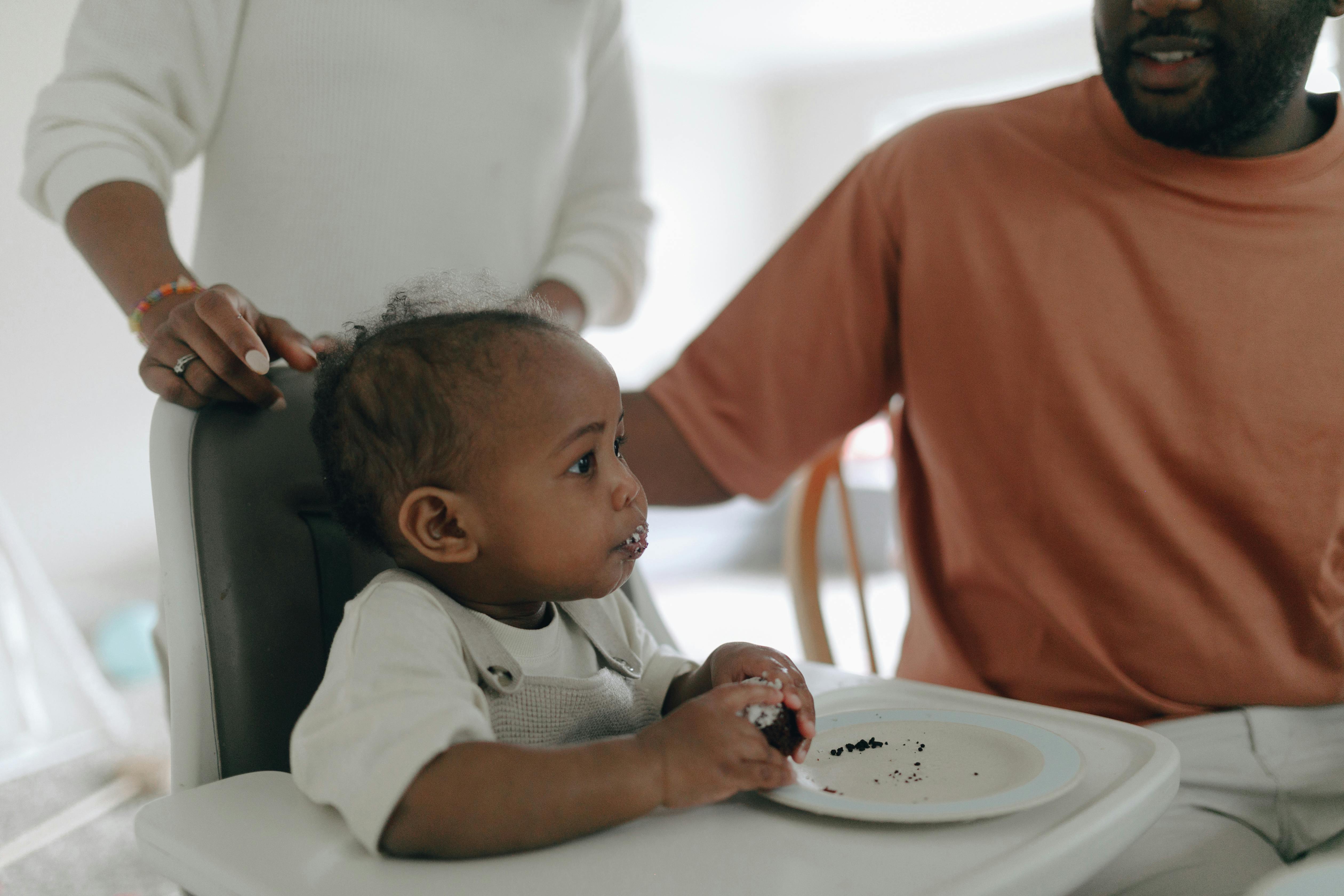 Black baby enjoying cake with family during first birthday celebration.