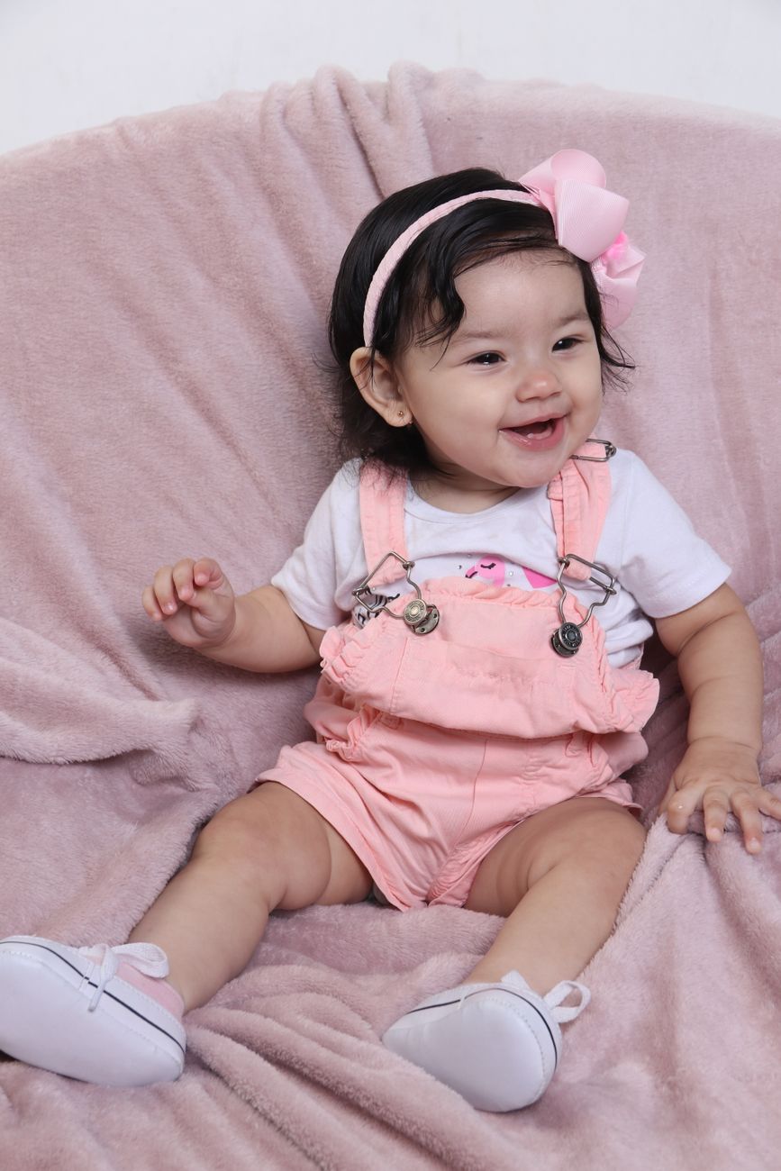 Bebê menina sentada e sorrindo, vestindo macacão rosa com laço