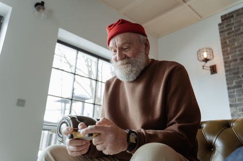 Free Senior man with beard and red hat playing a video game indoors, enjoying leisure time. Stock Photo