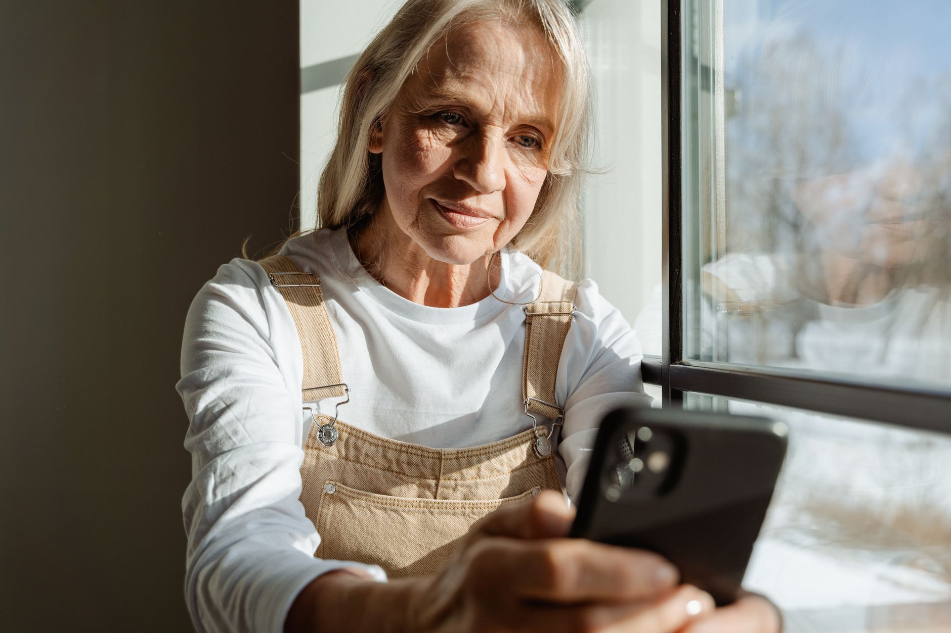 인천 어르신 스마트폰 무료 교육 7 A senior woman enjoys leisure time browsing on her smartphone by a sunlit window.