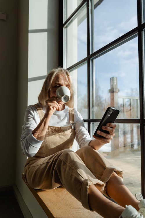 Woman in White Long Sleeve Shirt Drinking While Holding a Smartphone