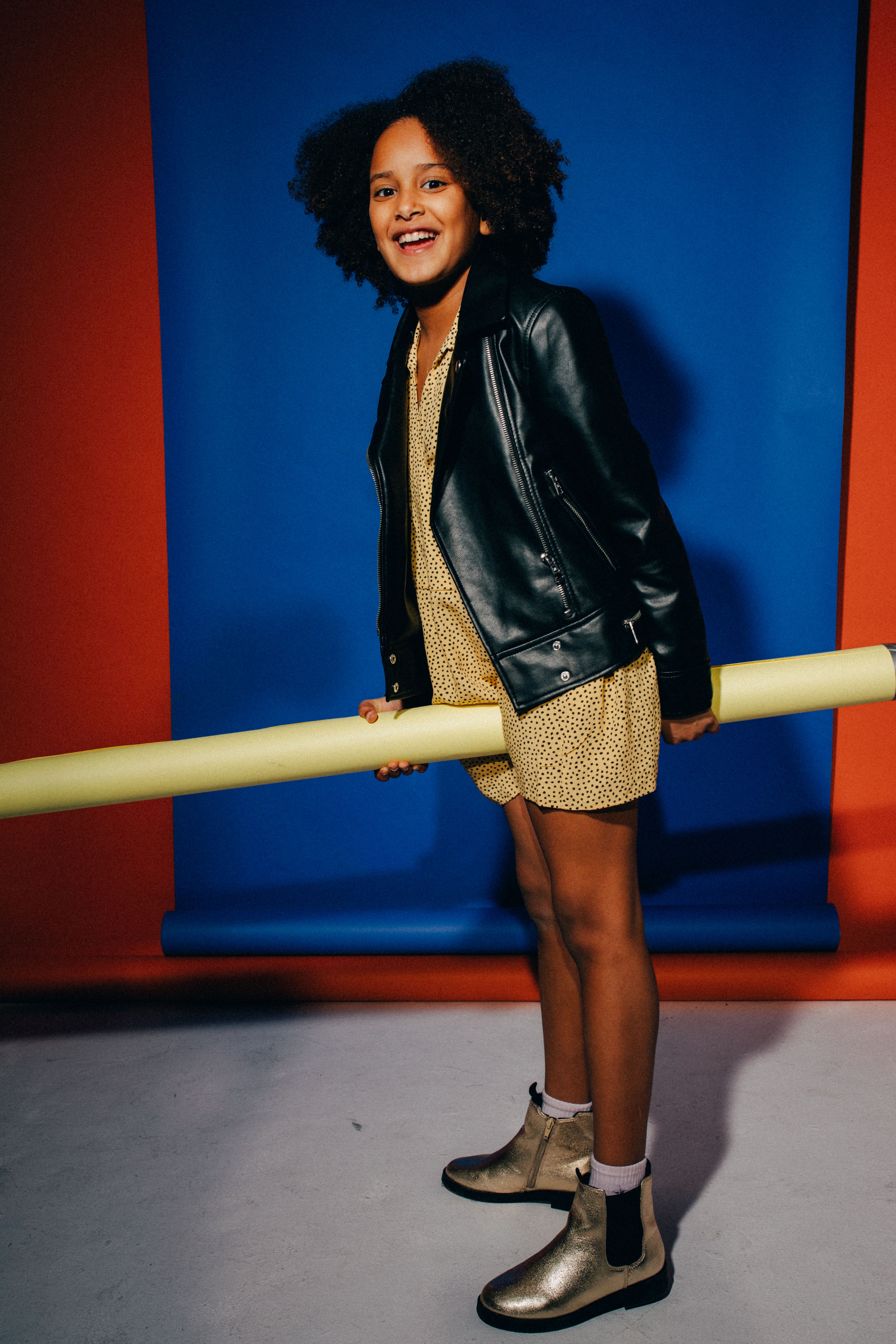 A cheerful young girl with an afro hairstyle holding a prop, showcasing stylish fashion indoors.