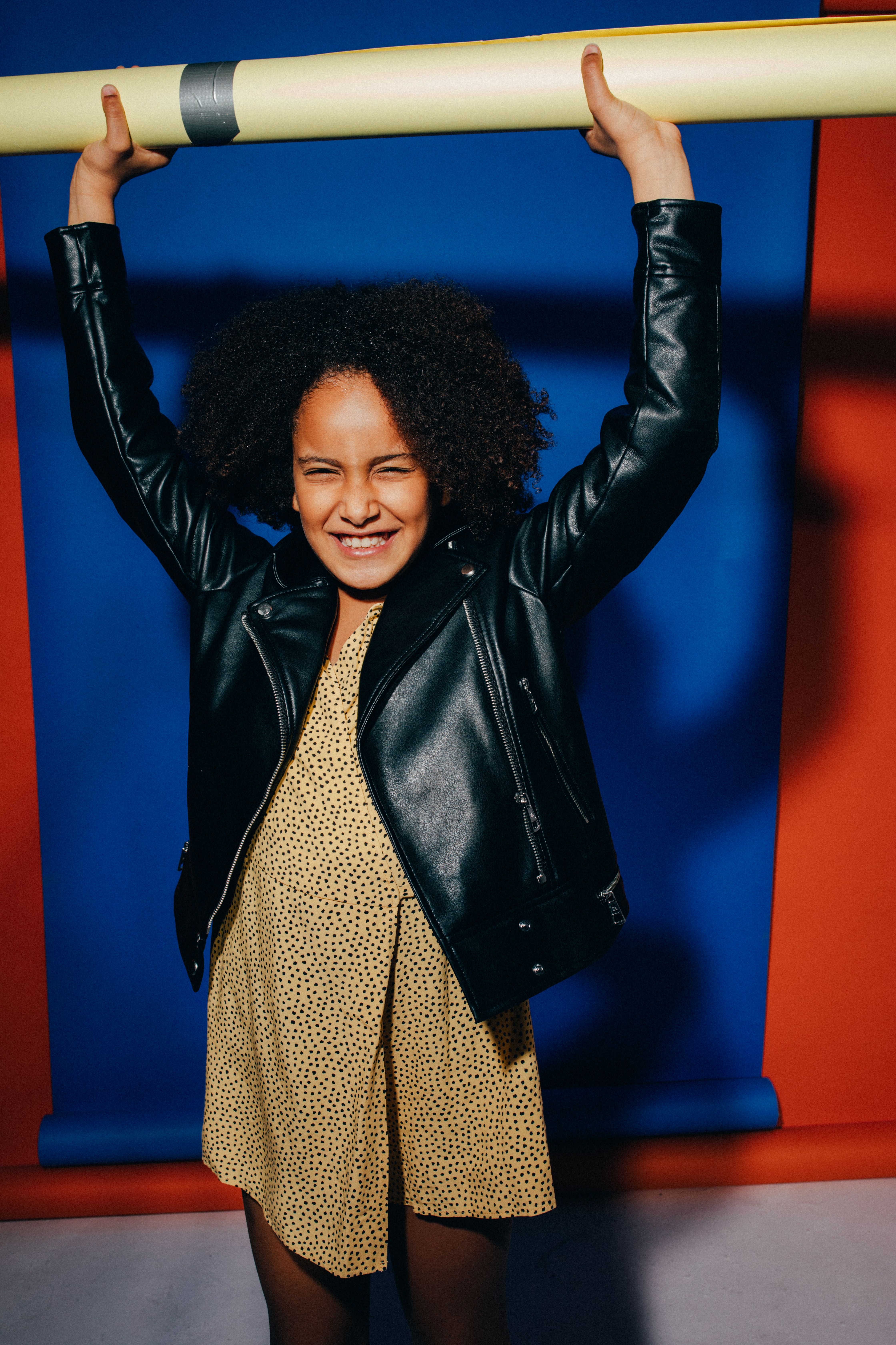 A smiling girl in a leather jacket raises a pole against a vibrant blue and red backdrop.