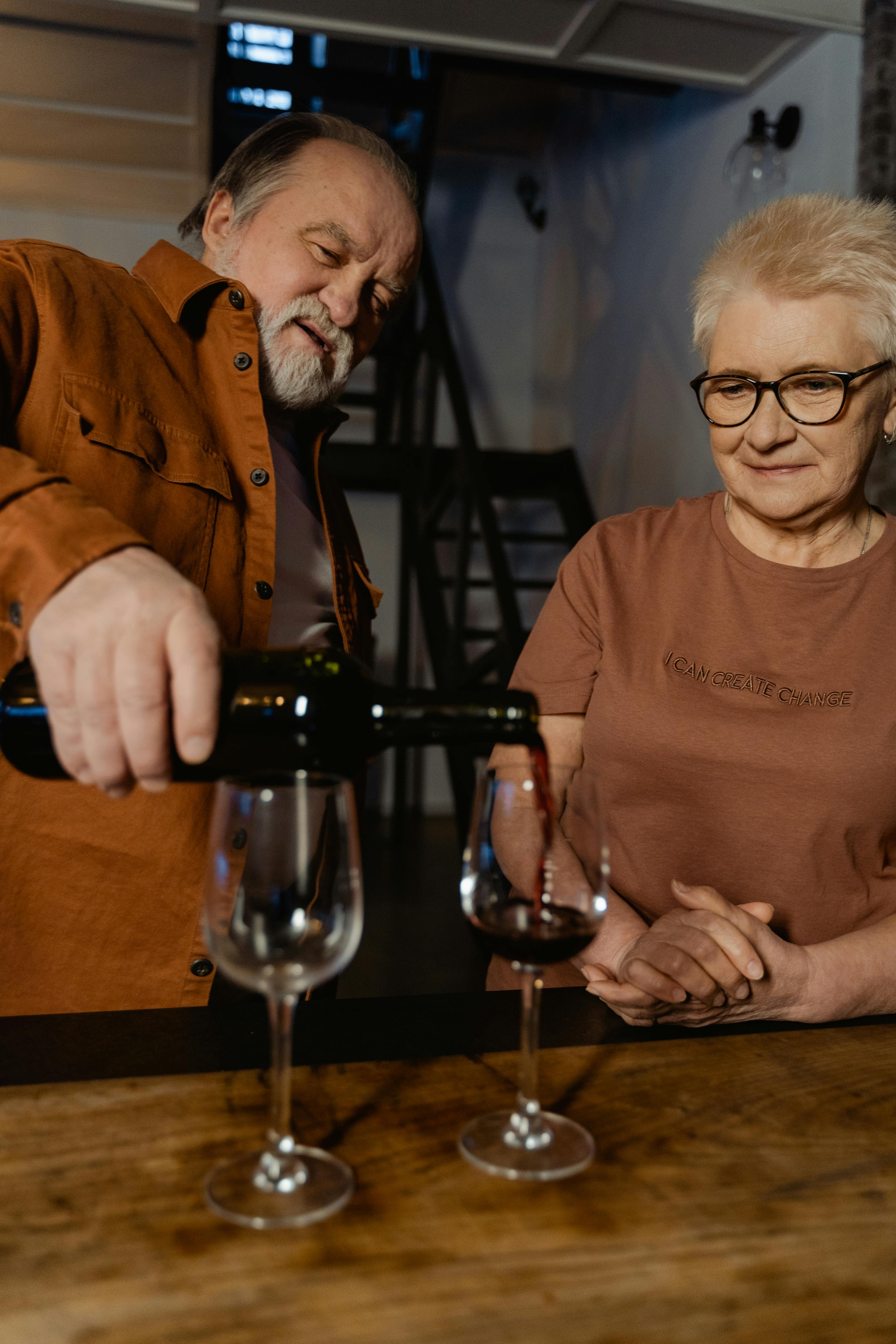Free An elderly couple enjoys a moment together pouring wine at home, showcasing warmth and togetherness. Stock Photo