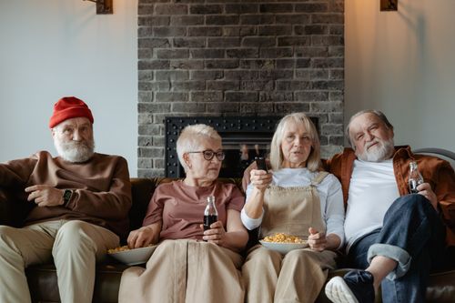 Free Four elderly friends enjoying a cozy TV time indoors with snacks and drinks. Stock Photo