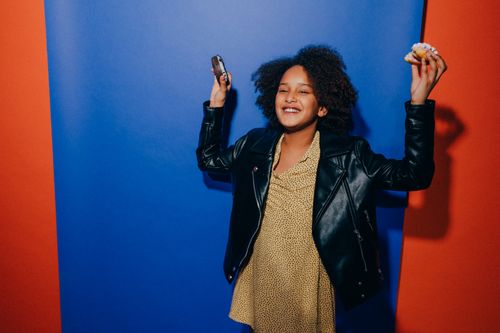 Free Happy girl with doughnut and phone smiles in stylish outfit against a vibrant, colorful backdrop. Stock Photo