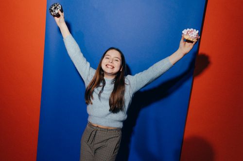 Free A smiling girl holding donuts, celebrating against a vibrant red and blue background. Stock Photo